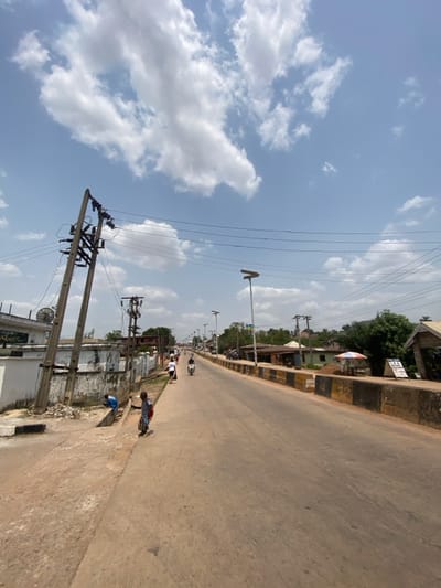 This image depicts a street scene in Afikpo, Nigeria, under a bright, partly cloudy sky. The perspective is from a low angle, looking down a two-lane, paved road that extends into the distance with a gentle incline.
In the left foreground, a tall, leaning wooden utility pole with multiple cross arms and numerous power lines dominates. Below it, two children are present on the unpaved shoulder. One child, wearing a blue top, is stooping near a concrete drain channel. The second child, in a light shirt and dark shorts, stands upright, facing the street while holding a red object, possibly a backpack. Behind the utility pole and children, a white building with a satellite dish on its roof is visible, fronted by a low concrete wall.
The paved road occupies the central and right portions of the frame. On the right side, a low concrete barrier, painted in alternating black and yellow/tan segments, separates the road from the adjacent area. A line of modern streetlights, each topped with a solar panel, runs alongside this barrier into the distance. Further down the road, several pedestrians walk on the left side, and a motorcycle carrying at least one individual travels away from the viewer.
On the right side of the road in the midground, various single-story structures or shops line the street. A white and red striped umbrella provides shade in front of one of these establishments, near a white sign displaying text, including "L.P.G." and "STATION." Green foliage from trees and bushes is visible behind these structures. The scene is illuminated by strong daylight, casting minimal shadows directly beneath objects.
