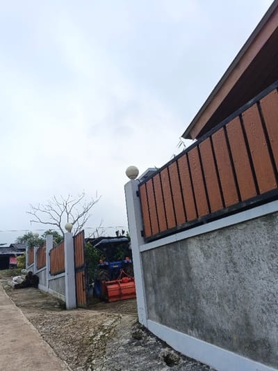 The image depicts an outdoor scene in Clacuc, Timor-Leste, under an overcast sky. In the foreground, a concrete pathway, partially covered with dirt and moss, extends from the lower left to the mid-ground. A light grey concrete wall with a white top trim runs along the right side, topped by a fence section composed of vertical brown wood-composite panels framed with black metal. A spherical white light fixture is affixed to a prominent fence post on the right. Another similar fence section and an open gate are visible further to the left, behind which a blue tractor, equipped with a red agricultural implement (likely a rototiller with "YANMAR" visible), is parked. The tractor is oriented with its front facing generally towards the viewer but angled slightly away. Behind the fence and left of the tractor, a bare tree with extensive branches stands against the sky. A pile of discarded materials or debris is present on the ground to the immediate left of the open gate. In the upper right corner, a segment of a building's brown-paneled roof is visible. Distant structures with dark roofs are partially observable in the background to the left. No individuals are present.