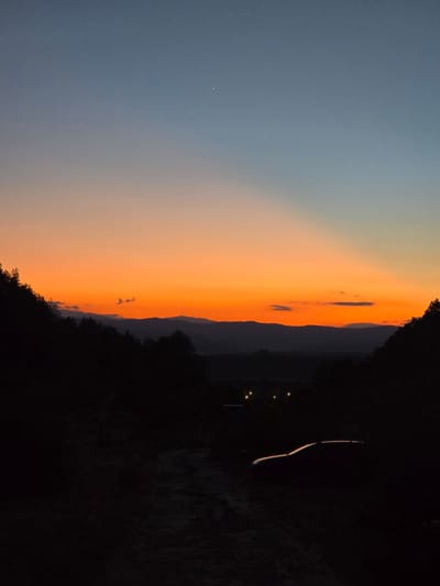 The image captures a sunset scene in Stob, Bulgaria, with the Rhodope Mountains in the background. The sky transitions from a deep blue at the top to vibrant orange and red hues near the horizon. Silhouetted trees frame the foreground, and a dirt road leads into the scene, with a car parked on the right side. Distant lights from a village are visible in the valley.