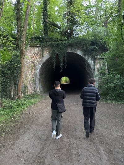 The image depicts an outdoor scene in Bures-sur-Yvette, France, showing two individuals on a dirt path approaching a stone-arched tunnel entrance. The primary subject is the tunnel, which appears dark inside, with a small, bright, circular opening visible at its far end, revealing green foliage and suggesting an exit.
One person is positioned closer to the camera, seen from behind, wearing a dark jacket (possibly black), dark trousers (resembling cargo pants), and white sneakers. They have short, dark hair and are looking towards the tunnel. A dark shoulder bag is slung over their shoulder. The second individual is further away, also facing the tunnel, dressed in a black puffer jacket, black trousers, and dark shoes.
The ground is a well-trodden dirt path, showing some dampness and small stones. The tunnel entrance is constructed from light-colored stone, partially covered in ivy and other green foliage. Dense trees and lush green undergrowth frame the path and the tunnel, creating a natural, somewhat wild setting. The lighting is soft and diffused, indicating an overcast day or shaded area, with brighter light visible at the tunnel's exit. The scene conveys a sense of exploration or passage through a natural landscape, likely a park or forest trail. There is no visible text in the image.