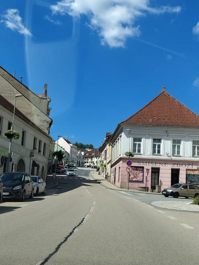 The image depicts a street scene in Volyně, Czechia, on a clear, sunny day with a bright blue sky and scattered white clouds. The perspective is from a vehicle traveling down a paved road that curves to the right.
On the left side of the street, a light-colored building with arched doorways and windows is visible, with plants in hanging baskets. Further down the street, other buildings with red-tiled roofs line the road. On the right side of the street, a pink building with white-framed windows features the signage "SMÍŠENÉ ZBOŽÍ" and "POTRAVINY". A pedestrian crossing is present in front of this building.
Several cars are parked along the street and some are in motion. A black Ford is parked on the left, and a silver station wagon is driving on the right. A few other vehicles are seen further up the road. The street is lined with trees and foliage in places. The overall impression is a typical European town street scene on a pleasant day.