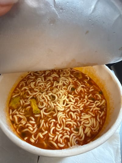 A close-up, overhead shot shows a cup of instant ramen noodles with a partially opened lid. The noodles are submerged in a reddish-orange broth, and some green specks and yellow pieces, likely vegetables or other ingredients, are visible. The cup is white and appears to be made of styrofoam. The background is out of focus, but suggests a light-colored surface, possibly a table with a napkin. No specific location in Qufu City, China is identifiable.