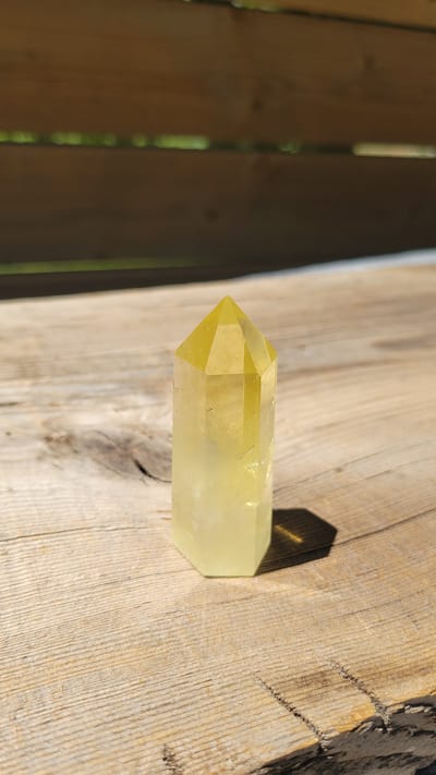 A single, faceted yellow crystal point stands upright on a weathered wooden surface. The crystal is translucent, with a lighter shade at the base and a deeper yellow at the pointed apex. Its hexagonal prism shape is clearly visible, with multiple facets meeting at the top. The wooden surface displays a rough texture, with visible grain lines and knots, and appears to be a piece of driftwood or a rustic tabletop. The background is out of focus, showing horizontal wooden planks with gaps through which glimpses of green foliage are visible. Sunlight casts a distinct shadow of the crystal onto the wooden surface to its right. The scene is outdoors, suggesting a natural or garden setting. There are no people or animals present in the image. The location in Terrebonne, Canada, is not discernible from the visual information provided.