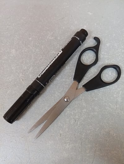 A black marker and a pair of black-handled scissors lie on a textured, light grey surface. The marker is positioned horizontally, angled slightly upwards from left to right, with its cap end on the left. Text is visible on the side of the marker. The scissors are placed diagonally to the right of the marker, with their blades pointing towards the bottom left of the image. The handles are open, revealing the metal pivot. The background surface appears to be a tabletop or workbench. There are no people or animals present. The lighting suggests an indoor setting. There is no information to suggest this image is related to Rezina, Moldova.