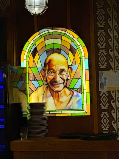 An indoor scene, likely a commercial establishment such as a restaurant, in Noumea, France, is depicted under subdued lighting. The central feature is a large, arched stained glass window portraying Mahatma Gandhi from the chest up, smiling and wearing round spectacles, against a multi-colored geometric background. Above the window, an industrial-style overhead light fixture with a wire cage emits a soft glow. Below the window, a wooden counter holds a tall stack of approximately 30 white plates, a single clear glass tumbler to its left, and two dark, shallow serving dishes to its right. To the far left, a reflective vertical surface, possibly a beverage cooler, emits a blue light. The wall to the right of the stained glass features dark paneling adorned with a repetitive, lighter-colored geometric pattern. A small, partially visible sign or poster, including a dartboard icon, is affixed to this patterned wall section. No individuals are present.