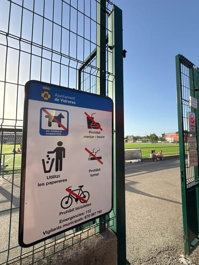 A close-up, eye-level shot captures a metal fence and a sign at the entrance to the Camp de Futbol Municipal in Vidreres, Spain. The sign, predominantly white with a blue border, displays several regulations in both Catalan and English, including prohibitions against domestic animals, eating and drinking, smoking, and cycling. It also instructs visitors to use the trash cans and provides emergency contact numbers. The sky is clear and blue. In the background, a well-maintained artificial turf soccer field is visible, with goalposts at either end. A few people are seated on a low wall near the field, and a scooter lies on the ground. Another sign on a green metal post to the right of the frame, also in Catalan, indicates "ACCÉS CAMP DE FUTBOL" and lists regulations, including "GOSSOS NO," meaning "dogs no."