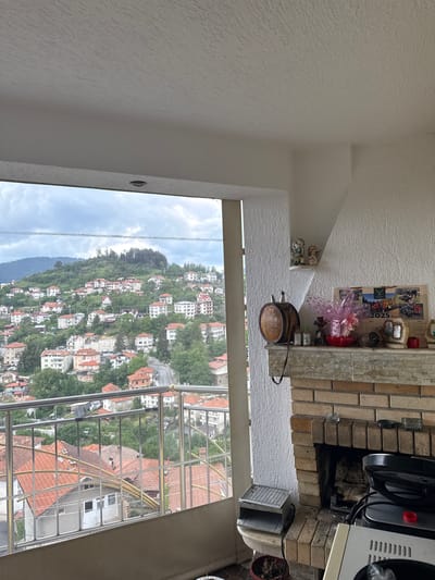 The image captures a detailed perspective from an elevated balcony or terrace, overlooking the village of Momchilovtsi, Bulgaria, on a cloudy day.

The left side frames a panoramic view of numerous multi-story houses with predominantly red or orange tiled roofs, densely distributed across steep, tree-covered hillsides. Winding roads are visible between the buildings, and distant mountains rise under an overcast sky crisscrossed by utility lines. A glass and polished metal railing with gold accents demarcates the balcony edge.

The right side reveals an interior corner featuring a prominent brick fireplace with a light-colored, textured stone mantelpiece. The firebox contains several logs. The mantelpiece displays various objects: a dark wooden barrel-shaped canteen with a leather strap, a red bowl containing an item wrapped in pink translucent fabric, several small decorative bottles, a black spherical object, and a 2025 wall calendar depicting agricultural machinery and outdoor scenes. Adjacent to the calendar are two small framed items and a small red box. Above the mantel, on a small white shelf, sit two ceramic figurines of cherubic figures. Below the mantel, an electric grill is placed on a small stand, and a dark potted plant with dried foliage rests on the floor. Partially visible in the lower right corner is a silver-toned appliance, likely a cooktop, with a black pan and a red cap on its surface. The interior walls are textured white plaster, and the white ceiling features a recessed light fixture above the balcony opening.