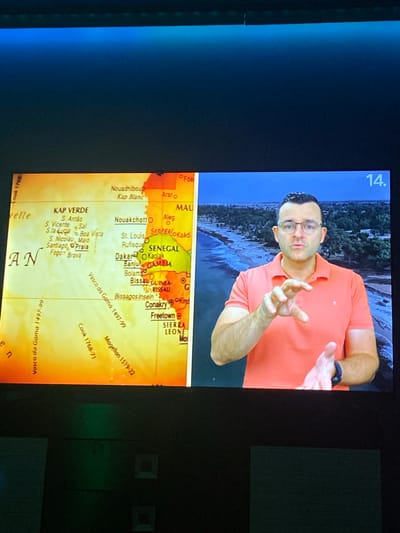 The image shows a split screen. The left side features a map of West Africa with names of cities and locations labeled. The right side shows a man wearing an orange polo shirt and glasses, gesturing with his hands in front of a backdrop of a coastline and ocean. The number "14." is visible in the upper right corner of the screen. The lower part of the image shows a dark surface that appears to be a wall with some square indentations. There is no information in the image that indicates the location is Benkovski, Bulgaria.