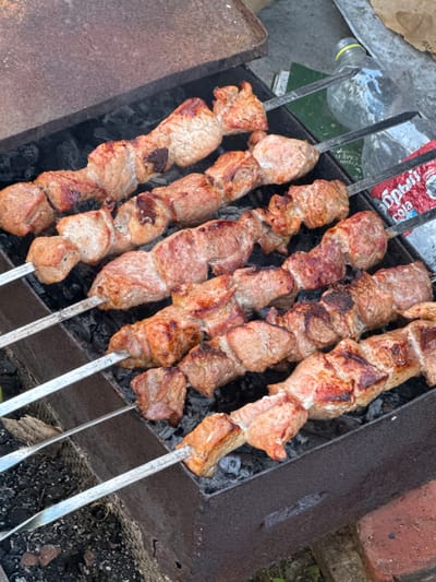 This is a close-up, overhead shot of meat skewers cooking over charcoal in a metal brazier. The skewers are arranged horizontally and fill most of the frame. The meat appears to be pork, cut into cubes, and is browned and slightly charred in places. The brazier is dark, rectangular, and shows signs of use. Scattered charcoal is visible beneath the skewers, some glowing red. In the background, to the right, a clear plastic bottle and a torn red and white wrapper with Cyrillic text are partially visible. The overall scene suggests an outdoor grilling activity. There are no people visible in the image. The location is identified as Bigila, Russia.