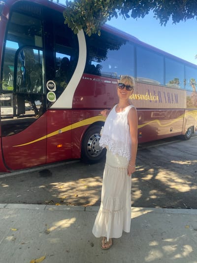 A woman stands on a sidewalk in front of a parked tour bus. The bus is maroon with a yellow stripe and text that reads "TRANSANDALUCIA". The woman is wearing a white lace top and a long, beige skirt. She has blonde hair and is wearing sunglasses. The background shows a clear blue sky and some trees. The location is Pol. Ind. La Palmosa, Spain.