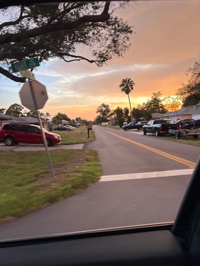 The image depicts a residential street in South Venice, United States of America, during sunset. The sky is filled with warm orange and yellow hues, with scattered clouds. A large oak tree with spreading branches is visible in the foreground on the left, partially obscuring a street sign and a stop sign.
On the street, several vehicles are parked. A red minivan is in the driveway of a house on the left. Further down the street, a blue pickup truck and a black pickup truck with a trailer are parked. Houses with typical suburban architecture are visible on both sides of the street. A tall palm tree stands out against the sunset sky. The road has yellow double lines marking the lanes. The image appears to be taken from inside a vehicle, as the bottom portion of the frame shows the interior of a car, including the dashboard and window frame. The overall scene conveys a peaceful, end-of-day atmosphere.