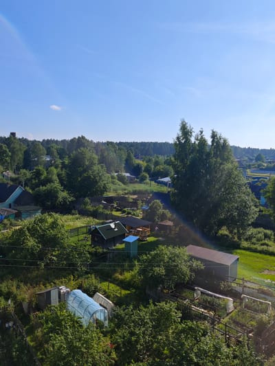 This is a medium shot, high-angle photograph of a rural Russian village scene on a sunny day. The sky is a clear, vibrant blue with a few wispy white clouds and a faint rainbow arching across the upper left. Below the sky, a dense, green forest surrounds a collection of small houses and outbuildings.
The scene appears to be a residential area with a mix of permanent structures and garden plots. In the foreground, to the left, are several greenhouses, one covered in a translucent blue material. To the right, there is a long, narrow building with a corrugated metal roof, likely a shed or workshop, and raised garden beds with protective coverings. A wooden fence encloses some of the garden areas.
In the middle ground, more houses with dark roofs are visible, nestled amongst the trees. A small, dark green building with a pitched roof and a blue structure next to it stands out. Further back, the forest continues, with patches of open green fields and more houses scattered throughout. A tall, slender smokestack or tower can be seen in the far distance, peeking above the treeline on the left.
The presence of greenhouses and garden plots suggests it is likely late spring or summer, a time for planting and growth in this location. The bright sunlight and clear sky indicate fair weather. The overall impression is one of a peaceful, working rural community. No people are clearly visible in the image, and there is no visible text. The location is identified as Zaklinye, Russia.