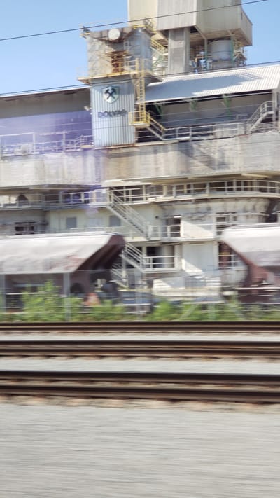 The image shows an industrial complex with a prominent logo bearing a hammer and crossed pickaxe above the word "DOLNÁ." The structure appears to be a factory or processing plant, with multiple levels, stairs, and railings. There are large, likely metal, structures and walkways. In the foreground, there are train tracks and a blurred landscape, suggesting the photo was taken from a moving vehicle, possibly a train. The sky is clear and blue. The provided context indicates this location is in Varín, Slovakia.