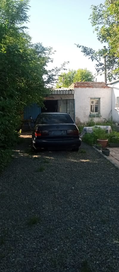 The image shows the rear of a dark-colored sedan parked on a gravel driveway in front of a partially open garage. The garage has a corrugated metal roof and a blue door. To the right of the garage is a white brick building with a single window. Green trees and foliage surround the scene, casting shadows. The sky is clear and blue, suggesting it is daytime, likely late afternoon or early evening due to the long shadows. There are several potted plants near the white building. The overall setting appears to be a rural or suburban area, possibly a backyard or a private property in Мариновка, Kazakhstan. No people are visible in the image, and the car appears to be stationary, indicating no particular activity is taking place other than the car being parked. The scene evokes a sense of quietness and everyday life. No visible text is present in the image.