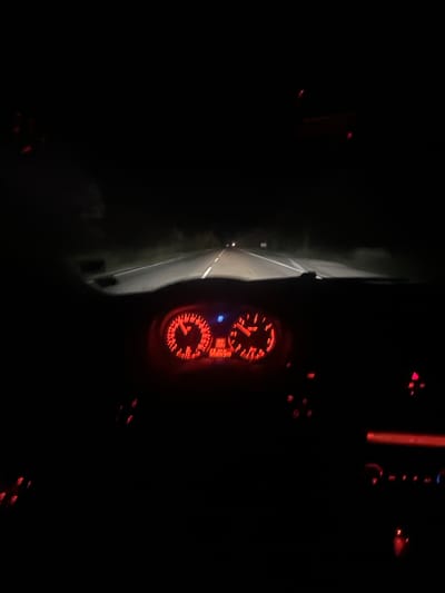 The image depicts the view from inside a vehicle driving on a dark road, possibly in Musachevo, Bulgaria. The dashboard gauges are illuminated in red, indicating the vehicle is in operation. The road ahead is dimly lit by the vehicle's headlights, revealing a dark asphalt surface with white lane markings. The darkness suggests a rural or unlit area. The car’s A pillar is partially visible to the left of the road.