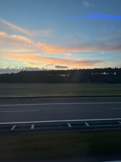 The image depicts a scenic view of a highway in Osogóra, Poland, during sunset. The sky is a canvas of soft pastel colors, with hues of pink, orange, and blue dominating the clouds. A dense forest lines the horizon, creating a dark silhouette against the vibrant sky. In the foreground, a highway stretches across the frame, with clear lane markings and a metal guardrail separating the lanes. The overall impression is one of tranquility and natural beauty, a peaceful moment captured on a journey through the Polish countryside.