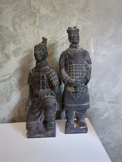 The image shows two terracotta warrior figurines displayed on a white surface against a textured, grey wall. The figurine on the left is in a kneeling position, while the one on the right stands upright. Both figures are dark in color and appear to be made of a material resembling clay or ceramic, with detailed armor and facial features. The lighting suggests an indoor setting. No specific location in the city of Taucha, Germany, can be identified from the image.
