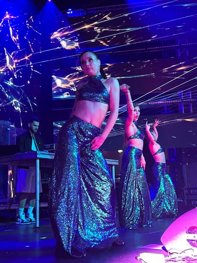 A group of female dancers perform on a brightly lit stage in Akhalubani, Georgia, wearing matching sparkly, teal-colored outfits. The dancers are positioned in a staggered formation, with the lead dancer in the foreground slightly to the left. The backdrop is a dynamic light show featuring abstract patterns in blue and orange hues, with thin, white lines crisscrossing the scene, creating a futuristic and energetic atmosphere. To the far left of the stage, a male DJ is visible behind a console, his attention focused on his equipment. The floor is a dark stage, illuminated by stage lighting that casts blue and pink glows. The overall scene depicts a lively performance with a focus on dance and visual effects.