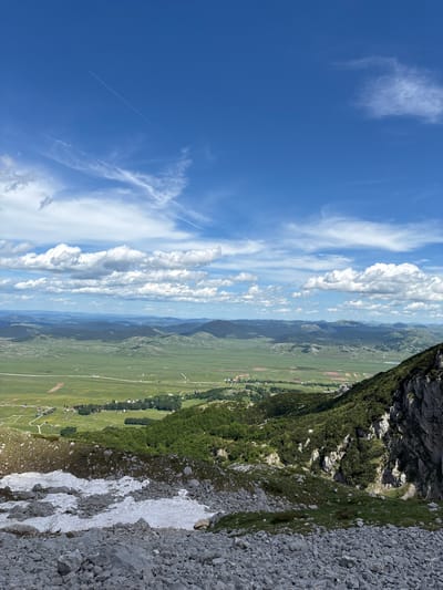 This is a wide-angle, high-angle view of a mountainous landscape under a bright blue sky with scattered white clouds and contrails. In the foreground, the terrain is rocky with patches of snow. The middle ground features a lush green valley dotted with trees and small buildings, suggesting a rural settlement or village. Further in the distance, rolling hills and mountains recede into a hazy horizon. The scene appears to be a natural landscape with human elements integrated into the valley.