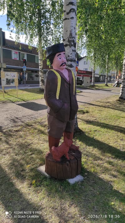 A wooden folk art statue of a man with a dark hat, brown jacket, and shorts, stands on a wooden log base in front of a birch tree. A small red dog sculpture is at the base of the log. The statue is located on a sidewalk in Iisalmi, Finland, with a street and buildings in the background. The buildings include shops with signs in Finnish, such as "Radio-outro Oy" and "Hautauspalvelu". The sky is blue and sunny with trees providing shade. The bottom of the image shows a watermark with "REDMI NOTE 9 PRO AI QUAD CAMERA" and a date "2026/7/8 18:36".