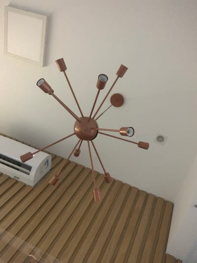 The image displays an interior architectural scene in Lauro de Freitas, Brazil, captured from a slightly upward angle. The central object is a large, multi-arm Sputnik-style chandelier, finished in a metallic copper or bronze, centrally mounted on a smooth, white ceiling. The chandelier consists of a spherical core from which numerous slender arms extend radially in various directions, each terminating in an empty cylindrical bulb socket.
To the upper left of the chandelier, a square, white-framed panel is affixed to the ceiling. Near the upper right, a small, circular recessed fixture, possibly a vent or light, is visible in the ceiling. Adjacent to the chandelier's main attachment point, a smaller, circular base matching the chandelier's finish is also present on the ceiling.
In the lower left quadrant, a white, wall-mounted split-system air conditioning unit with a digital display is installed. Below the air conditioning unit and extending across the lower portion of the image is a wall or partition clad with uniformly spaced vertical wooden slats, presenting a textured surface. No individuals, actions, or interactions are present in the depicted environment.