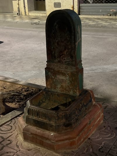 The image displays an outdoor scene in Alcàsser, Spain, centered on a freestanding, heavily corroded drinking fountain. The fountain, constructed of metal, primarily cast iron, exhibits extensive surface rust in various shades of brown and orange, with remnants of dark green paint visible on its column. Its design features a tall, slender column with an arched top, containing an obscured embossed motif. Below the column is a hexagonal basin, currently dry and containing accumulated organic debris. The fountain rests on an octagonal base composed of reddish, brick-like material, which in turn sits on dark reddish-brown sidewalk tiles adorned with a repeating curvilinear pattern. To the immediate left of the fountain, an exposed dirt patch, possibly a tree pit, is filled with dry leaves and debris, with a flat metal bar partially visible within it. A grey asphalt road stretches into the background. On the left side of the road, a light-colored building features two vertical utility pipes and a section of a checkered sidewalk. On the right, another building presents a closed dark grey corrugated metal security shutter, also with a segment of checkered sidewalk in front. The scene is illuminated by artificial light, suggesting nighttime or low-light conditions. No individuals, animals, or actions are depicted within the frame.