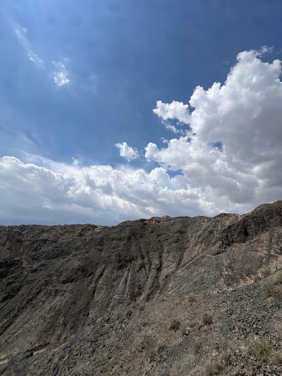 The image shows a desolate, rocky mountain landscape under a blue sky with white clouds. The foreground is dominated by dark gray, jagged rocks and scree, with sparse, dry vegetation. The terrain slopes upwards and recedes into the distance, forming the rugged side of a mountain. In the upper portion of the image, the sky is filled with various cloud formations, ranging from wispy streaks to large, puffy cumulus clouds. The scene evokes a sense of natural ruggedness and vastness, typical of arid or mountainous regions.