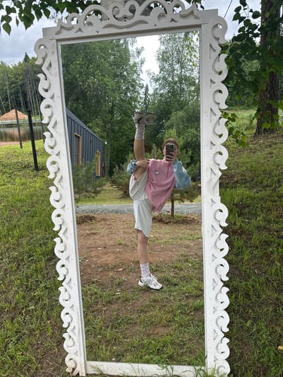 The image depicts a full-length mirror with an ornate white frame, placed outdoors in a grassy area. A young woman is visible in the reflection, performing a high leg split. She is wearing a pink and blue sweatshirt, white shorts, and white athletic socks and shoes. She is holding her phone to take the picture. In the background, a modern-style dark building, trees, a body of water, and a hill with a pile of sand are visible. The location is identified as Novaya Vozhoika, Russia. The weather appears overcast.