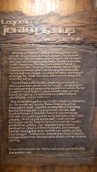This image depicts a vertically oriented, rectangular wooden plaque, carved with relief text and decorative elements, located in Glantengan, Indonesia. The plaque, presented indoors against a lighter wood-paneled wall, is illuminated from above.
The top section of the plaque features "Legenda Jenang Kudus" (The Legend of Kudus Jenang) in stylized, raised characters, with "Syekh Jangkung (Saridin)" carved below in a simpler font. The main body of the plaque contains multiple paragraphs of carved Indonesian text. This text details the legend of Jenang Kudus, originating in Desa Kaliputu, Kecamatan Kota, Kabupaten Kudus, Jawa Tengah.
The narrative describes an incident involving Mbah Depok and his grandchild, who fell into a river. Historical figures mentioned include Sunan Kudus and Syekh Jangkung (Saridin), who intervened in the panic. The legend attributes the revival of the child, who was only "mati suri" (clinically dead, but revived), to the creation of "Jenang Bubur Gamping" using rice flour, salt, and coconut milk. The text concludes by explaining that this legend is the basis for the annual "Kirab Tebokan" or "Arak-arakan Jenang" celebration held on 1 Muharram in Desa Kaliputu, as an expression of gratitude for the blessings derived from the Jenang business. The information on the plaque is credited to the Dinas Kebudayaan dan Pariwisata Kabupaten Kudus (Department of Culture and Tourism of Kudus Regency) and other sources. No individuals are visible in the scene; the focus is solely on the informational plaque and its content. The environment suggests a public indoor display setting.