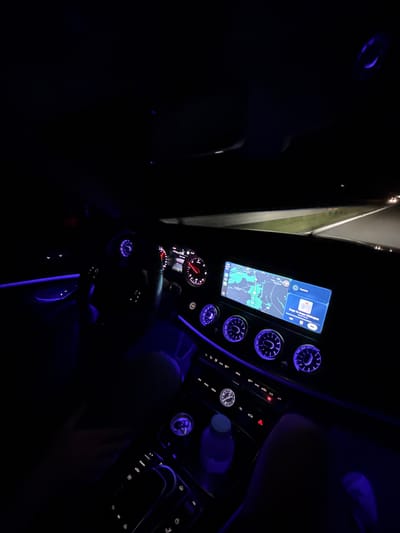 The image shows the interior of a car at night. The dashboard is visible, featuring a navigation screen displaying a map, illuminated vents, and various controls. The car appears to be stationary, and part of a road can be seen through the windshield. The navigation screen shows Церово, Bulgaria, indicating the location. There are no people visible in the image.