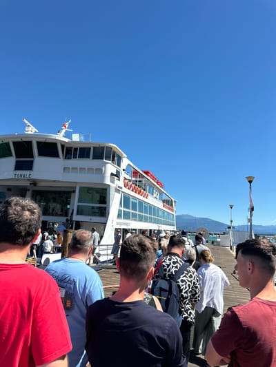 A group of people are boarding a white ferry named "TONALE" on a sunny day in Desenzano del Garda, Italy. The ferry is docked at a pier, and the people are lined up, waiting to board. The sky is clear and blue, and the water is calm. In the background, mountains can be seen. The people are dressed in casual clothing, and some are carrying bags. The atmosphere is relaxed and orderly.