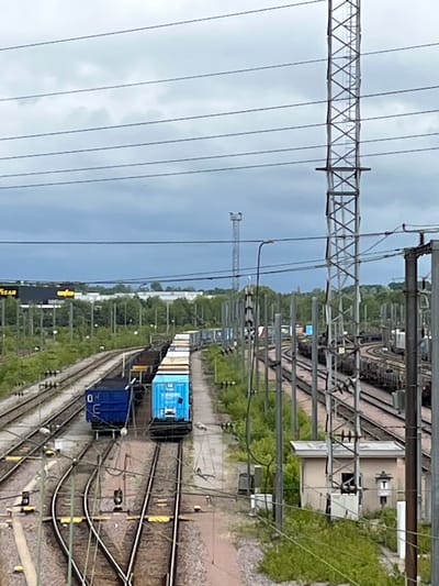 The image depicts a railyard in Dudelange, Luxembourg, under a cloudy sky. Several sets of train tracks are visible, with multiple freight trains on them. One train in the foreground consists of at least two blue cargo containers and several white-topped containers. Another train further back is composed of numerous dark-colored cargo wagons. Overhead power lines crisscross the scene, supported by tall metal pylons, including a prominent lattice tower on the right. Vegetation, including trees and bushes, lines the sides of the tracks. In the background, industrial buildings with yellow signage, possibly from a commercial area, are visible. A small, light-colored utility building with a window and a sign featuring a stylized character is situated near the tracks on the right side of the image. There are no people visible in the frame.