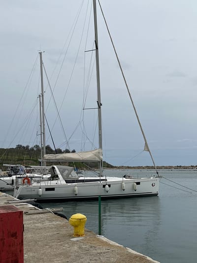 A white "Oceanis 48" sailboat is moored in the harbor of Limenas Avdiron, Greece, under an overcast sky. The sailboat, positioned parallel to a concrete pier on its port side, features two tall masts with extensive rigging, a furled main sail covered in beige canvas, and a grey dodger over the cockpit. Multiple white cylindrical fenders are suspended along its hull. A distinctive dark logo is visible on the bow. Mooring lines extend from the vessel to unseen points and potentially to the pier. In the foreground, a yellow mooring cleat is prominently affixed to the concrete pier, with a partial view of a red structure to its left. The calm water reflects the grey sky. The background reveals a verdant landmass with trees to the left and a rocky breakwater protecting the harbor to the right. No individuals are present in the scene.