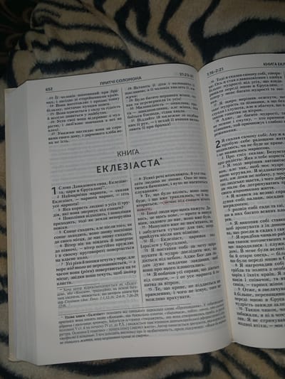 This is a close-up, top-down, indoor shot of an open book, likely a Bible, resting on a dark, textured surface. The book is open to two pages of text. The left page contains a heading "ПРІТЧІ СОЛОМОНА" and text in Ukrainian, with numbered verses. The right page has a heading "КНИГА ЕКЛ" and text in Ukrainian, also with numbered verses. The lighting is dim, creating shadows and highlighting the texture of the book's pages and the surface it lies on. There are no people or any indication of location in Nekrashi, Ukraine.