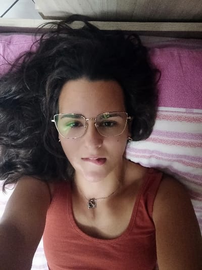 A person lies on a striped purple and white bed, hair spread across the pillow. They wear a reddish-orange top and a silver necklace with a pendant. Glasses perch on their face, reflecting green light. They gaze toward the camera with a slight smile. Part of a wooden headboard is visible on the right. The person's arm is raised, partially obscuring the top left of the frame.
