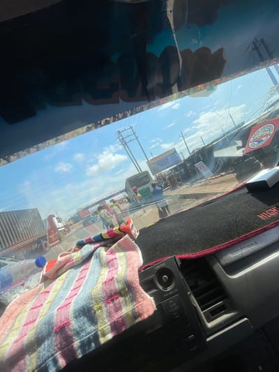 This media file captures a view from inside a vehicle looking out through the windshield and side window. The scene outside appears to be a bustling street in Mowe, Nigeria, likely during daytime with a clear, blue sky and scattered clouds, indicating fair weather.
The windshield reflects the sky and shows a busy street scene with numerous vehicles, including trucks, cars, and possibly some motorcycles. People are visible on the roadside, suggesting commercial activity or daily life. There are buildings and power lines, typical of an urban or semi-urban environment. Some signs and posters are visible on the street, though the text is largely illegible due to the image quality and distance. One partially visible circular sticker on the dashboard or windshield reads "TERMINAL ONE".
Inside the vehicle, a colorful striped cloth is draped over what appears to be part of the dashboard or interior. A plastic water bottle with a blue cap is also visible in the foreground, placed near the cloth. The interior of the vehicle includes visible car parts like an air vent and a car stereo system with buttons and labels, including "TUNE FILE" and "PUSH AUDIO CTRL".
The overall impression is a snapshot of everyday life and travel in a busy Nigerian town, viewed from the perspective of a vehicle occupant. The bright sunlight suggests it's likely morning or afternoon.