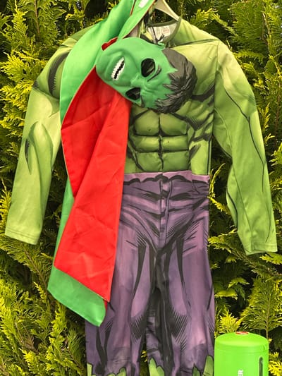 A Hulk-themed costume is prominently displayed against a dense, vibrant green foliage background, likely a coniferous hedge or bush, under natural daylight. The costume consists of a green, long-sleeved jumpsuit top designed with printed muscle definition (pectorals, abdominals, and arm muscles) in shades of green and black linework. A white clothing hanger is visible at the top, supporting the costume. On the inside collar of the jumpsuit, a small white tag with the "MARVEL" logo is discernible.

Draped over the left shoulder and arm of the costume is a red cape, which is lined with a contrasting green fabric. A green plastic mask, designed to resemble the Hulk's face with black hair details, eyeholes, and white painted teeth, rests on the upper torso of the green jumpsuit. Below the jumpsuit top, a pair of purple pants, also part of the costume, feature printed details mimicking torn fabric and muscular legs in purple and black linework. In the bottom right corner of the frame, a small, cylindrical bright green container with a red lid is partially visible, displaying the letters "ER" on its side. The foliage behind the costume is uniformly green with textured, needle-like leaves, suggesting an outdoor setting.