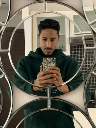 A male individual is depicted taking a selfie in a segmented, decorative mirror, reportedly in Ksar El Majaz, Morocco. The subject has dark, curly hair, a trimmed mustache, and a chin beard. He is wearing black sunglasses perched on his head, a dark green hoodie, and a black turtleneck shirt. He holds a smartphone vertically with both hands, positioned in front of his chest, to capture the reflection. The smartphone has a light-colored, patterned case. Mirrored text on the hoodie's chest is partially visible, indicating "BOSTON" and letters resembling "TICS". The mirror's frame is silver-toned, composed of interconnected circular and semi-circular segments. The reflected background reveals an indoor setting: a light-colored wall or cabinet is visible to the viewer's right of the subject, and to the viewer's left, white cabinetry is positioned above a section of dark, small-square tiling, consistent with a kitchen or bathroom backsplash.