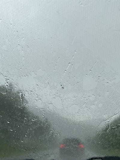 The image shows a view through a wet car windshield, looking out at a hazy, overcast day. The windshield is covered in raindrops and streaks, distorting the view of the outside. In the distance, the taillights of another car are visible, indicating that the vehicles are on a road, possibly in a rural or suburban area. The surrounding environment appears to be green, suggesting trees or vegetation, though the low visibility makes it difficult to discern fine details. The overall impression is one of a dreary, rainy day. There is no indication of the city of Кулмас, Russia, within the image.