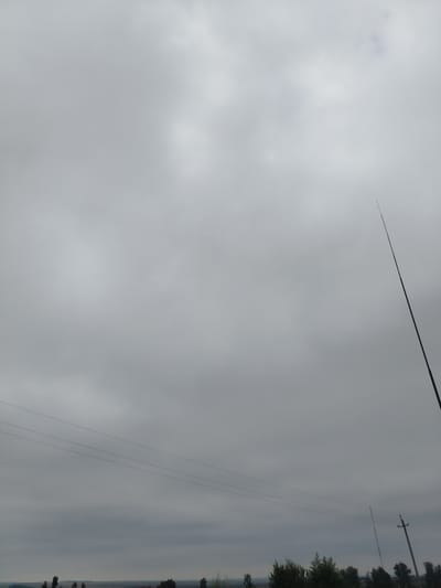 The image depicts an overcast sky with diffused light, suggesting a cloudy or hazy atmosphere. In the foreground, silhouetted against the sky, is a thin, dark, vertical pole extending from the bottom edge of the frame towards the upper right. This pole appears to be a man-made structure, possibly an antenna or a part of a utility pole.
Further down, a series of thin, dark lines traverse the frame horizontally, indicating power lines suspended between unseen supports. Below these lines, the horizon is visible, featuring a dark band of trees and vegetation. The landscape appears flat, with a few indistinct structures or features visible in the distance.
The overall impression is a rather bleak or muted scene, dominated by the sky and the utilitarian elements of the landscape. There are no people or animals visible in the frame, and no discernible actions or interactions taking place. The environment suggests an outdoor, rural, or semi-rural setting, likely in the town of Stepnoe, Russia, given the context.