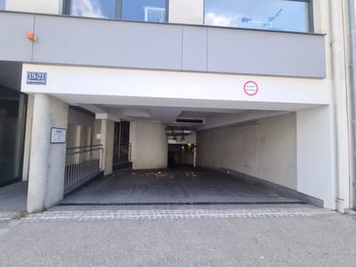 The image shows the entrance to a parking garage. A sign with "19-21 Hauptstraße" is visible on the wall above the entrance, indicating the address. A red circular sign with "Ausfahrt freihalten" is also visible, meaning "Keep exit clear." The parking garage has a ramp leading down into it, and the ground appears to be wet in places. There are some debris on the floor of the garage. The lighting suggests it is daytime, and the weather appears to be overcast. The scene is an urban setting, likely in Mödling, Austria. There are no people visible in the image. A bicycle is visible through a window on the upper floor of the building.