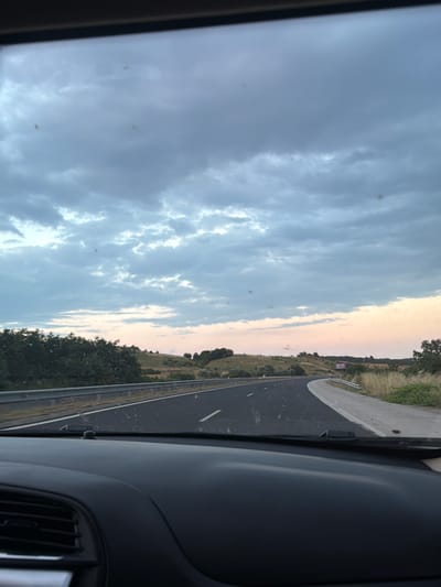 The image shows a view from inside a car traveling on a highway. The road ahead curves to the right, bordered by a metal guardrail and dry vegetation. Rolling hills with scattered trees and bushes form the background, under a cloudy sky with streaks of pink and orange from what appears to be a sunset. There are some dark spots on the windshield, possibly dirt or water marks. The interior of the car is visible in the foreground, including the dashboard and an air vent. A purple billboard is visible in the distance on the right side of the road.