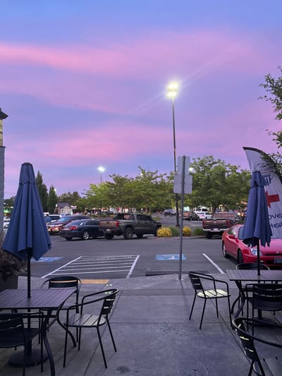 A parking lot scene at dusk with a vivid, pink and blue sky. Foreground features an outdoor patio with dark metal tables and chairs, sheltered by blue umbrellas. Cars are parked in marked spaces in the middle ground. Streetlights illuminate the parking area, casting a glow on the trees lining the lot. A red car is parked at the very edge of the frame. A sign is seen on the left of the frame.