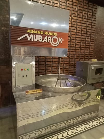 The image shows a large, stainless steel mixing bowl, a metal control panel with green and red lights, and a sign that reads "Mesin Mixer Pengolahan Jenang" ("Mixer machine for processing jenang"). The mixing bowl is attached to a metal frame and has a set of mixing arms inside. The control panel is located on the side of the bowl, and the sign is below it. The background is a tiled wall, with a brown and white color scheme. There are no people present. This scene appears to be inside a factory or kitchen at the coordinates -6.809, 110.843, which are located in the city of Kudus, Indonesia. The scene is likely part of a factory or facility for the production of "jenang", a traditional Indonesian dessert.