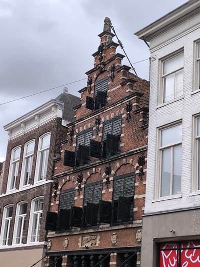 The image shows a close-up view of the facades of several historic buildings in Haarlem, Netherlands, under a cloudy sky. The central building is constructed of red brick with a stepped gable roof, adorned with decorative black ironwork and a statue at the apex. It features multiple rows of arched windows with black shutters, some of which are open. To its left is a building with a lighter colored brick facade and white trim, also with arched windows. To the right, a white brick building with a flat roof is visible, featuring large rectangular windows. The buildings appear to be shops or residences situated on a street, as indicated by the lower portions of the structures, which show entranceways and what might be shop windows. The overall impression is one of old-world architecture in an urban setting.
