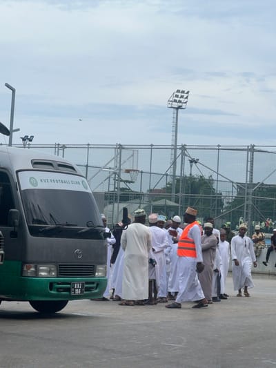 The image displays an outdoor scene in Zanzibar City, Tanzania, featuring a dark grey and green minibus alongside a group of individuals on a paved area. The minibus, positioned on the left, has a white banner across its windshield stating "KVZ FOOTBALL CLUB" with a circular emblem. Its front license plate reads "KVZ 156". Approximately 15 to 20 adult males are present, mostly wearing white long robes (kanzus) and traditional embroidered caps (kufis). One man in the foreground is walking towards the left, distinguished by a bright orange reflective safety vest worn over his white robe. Other individuals are standing in small groups, some appearing to converse. The background features a chain-link fence enclosing a sports facility, identifiable by a visible basketball hoop and a tall floodlight tower with multiple light fixtures. Green vegetation is visible beyond the fence, and the sky is overcast with light grey clouds. A small, dark shape, possibly a bird or a distant aircraft, is visible in the upper sky.