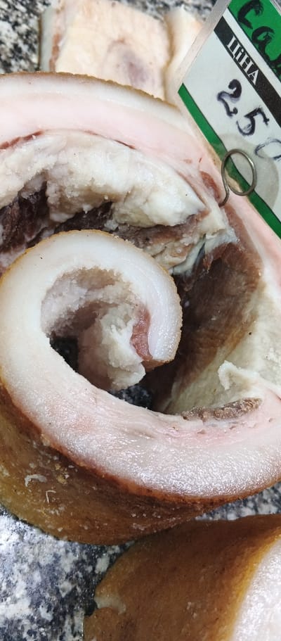 A close-up, overhead shot shows slices of raw pork belly. The meat is pale pinkish-white with distinct layers of fat. Some of the slices are rolled, revealing a spiral pattern. A small white tag with green lettering and the number "25" written in black marker is attached to the meat with a metal ring. The background consists of a marbled countertop. The image is likely from a butcher shop or market in Ploske, Ukraine.