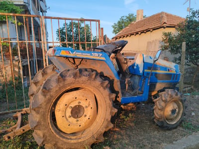 The image depicts a blue and white tractor with "LAND HOPE 197" and "ISEKI" branding, parked outdoors in what appears to be a rural or semi-rural setting. The tractor is positioned on dry grass and dirt, with a rusty metal fence in the foreground. Behind the tractor, a light-colored building with a tiled roof is visible, along with trees and vegetation. The sky above is clear and blue. The scene is illuminated by natural sunlight, casting shadows and highlighting the textures of the tractor's tires and the surrounding environment. The overall impression is one of a static, still outdoor scene. The location is identified as Bryastovets, Bulgaria, suggesting a specific geographical context for the image.