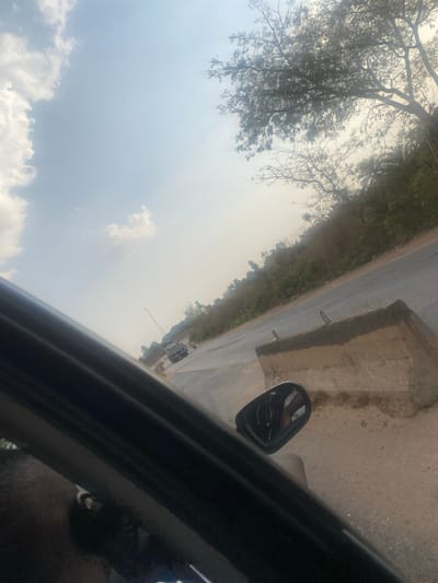 A tilted view from inside a car shows a road bordered by vegetation and concrete barriers. A side mirror reflects the roadside. In the distance, vehicles and a crane are visible. The sky, filled with clouds, occupies a significant portion of the frame, dominating the upper-left area. The car's interior and the person are partially visible.