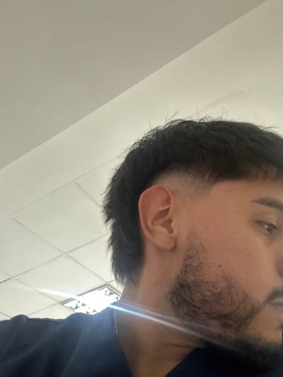 The image displays the right side of a male individual, visible from the mid-chest upward, against an indoor background. The subject has dark hair, styled with a high fade on the sides that blends into longer hair on the top and at the nape. A dark, trimmed beard covers his jawline and connects to his sideburns. His right ear is fully visible. He is wearing a dark blue collared garment, and a thin silver-colored chain is visible around his neck. His right eye, brow, and a portion of his nose and upper lip are discernible, with his gaze directed slightly downward and away from the camera's perspective. The background features a light-colored, possibly white or off-white, drop ceiling with distinct square panels. A bright light fixture, likely fluorescent, is partially visible in the upper left background, casting a noticeable lens flare across the lower left quadrant of the image, notably over the subject's neck and shirt. The environment provides no specific visual cues to determine a precise location within the city of Adrogué, Argentina.